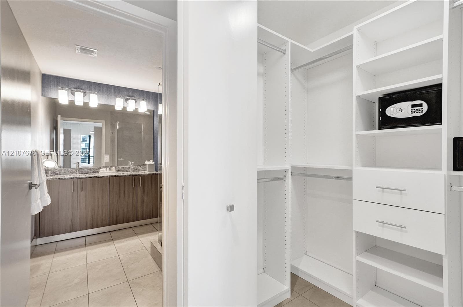 Walk-in closet featuring white built-in shelving, drawers, and hanging rods, accompanied by a bathroom with a dual-sink vanity, stone-finish countertops, tile flooring, and wall-mounted light fixtures