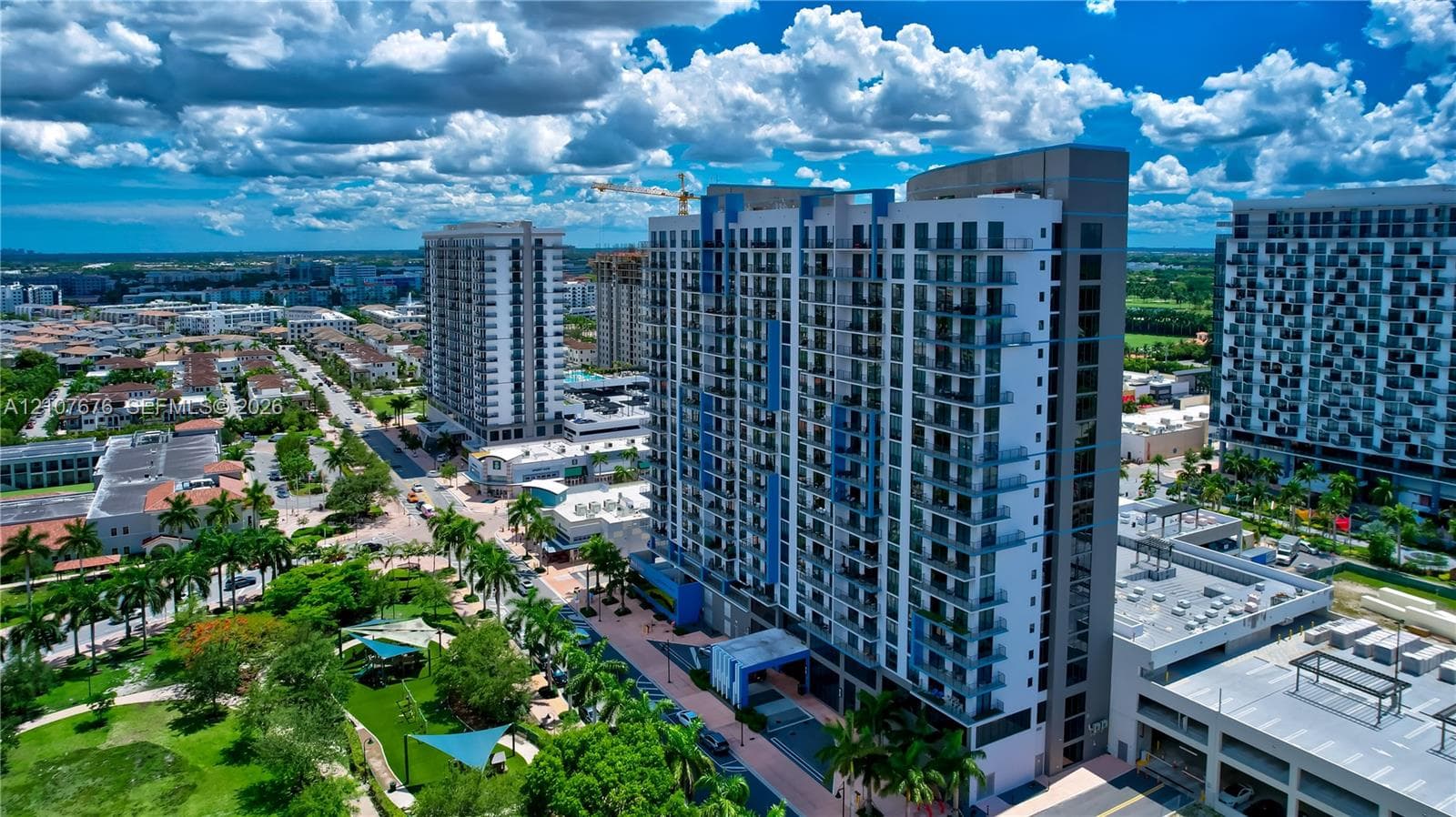 High-rise architectural structure featuring a grid glass facade and individual balconies, situated in a dense urban environment with surrounding multi-story developments and paved pedestrian access