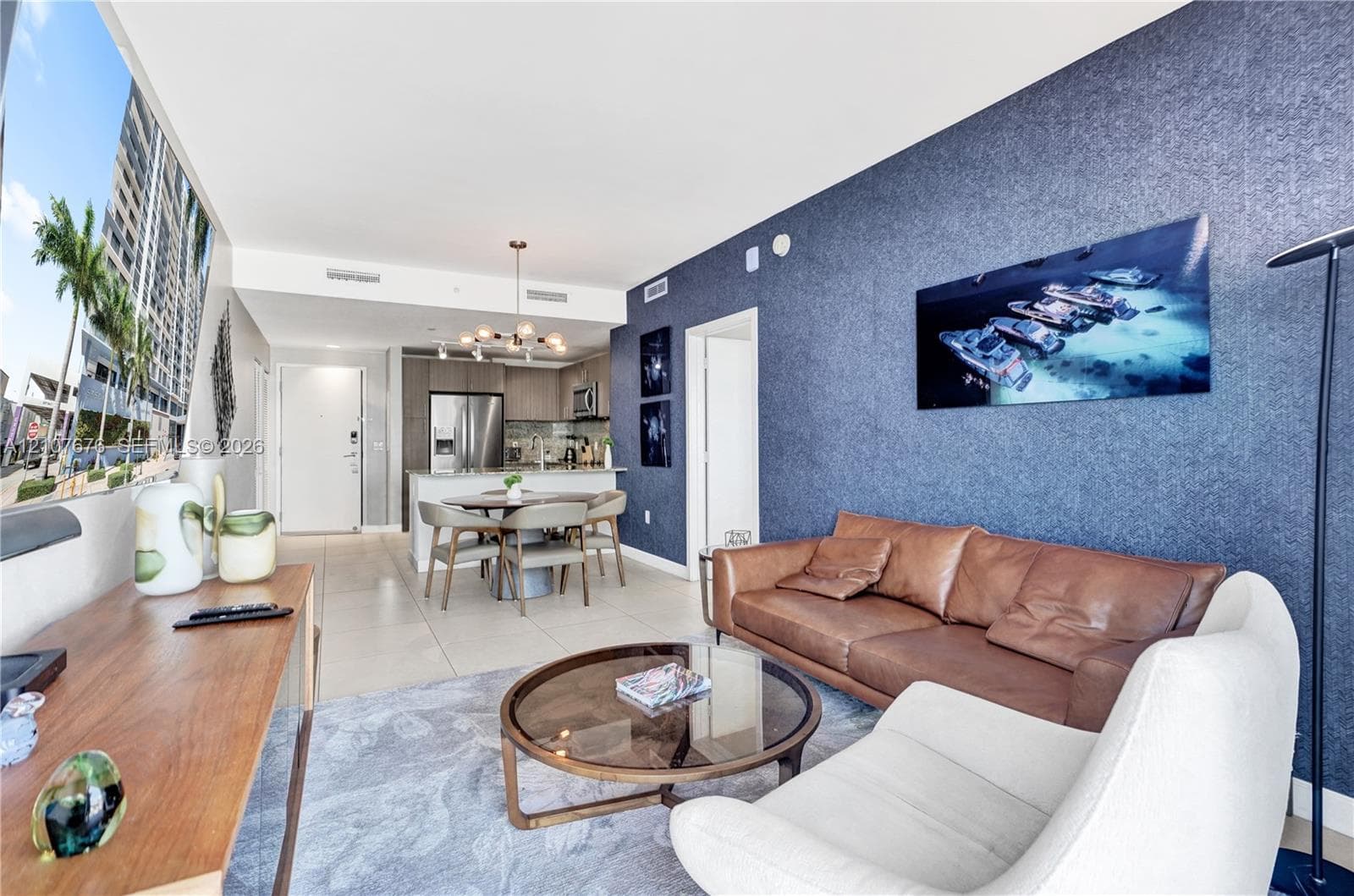 Open-concept living space featuring large-format tile flooring, a prominent textured navy accent wall, stainless steel kitchen appliances, and a contemporary multi-light chandelier