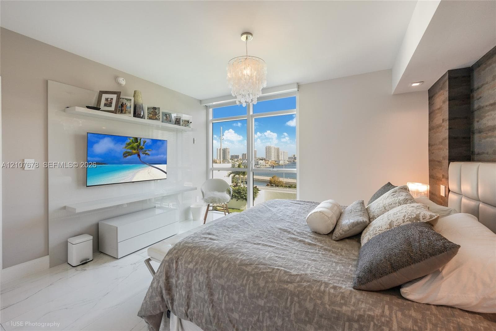 Floor-to-ceiling windows, custom built-in white media console with integrated shelving, wood-finish accent wall, polished stone-finish flooring, and decorative chandelier