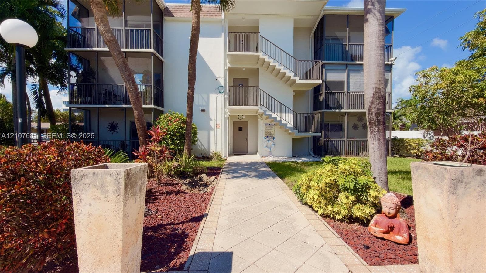 Three-story residential building exterior featuring screened-in lanais, external staircase, concrete paver walkway, and mature tropical landscaping