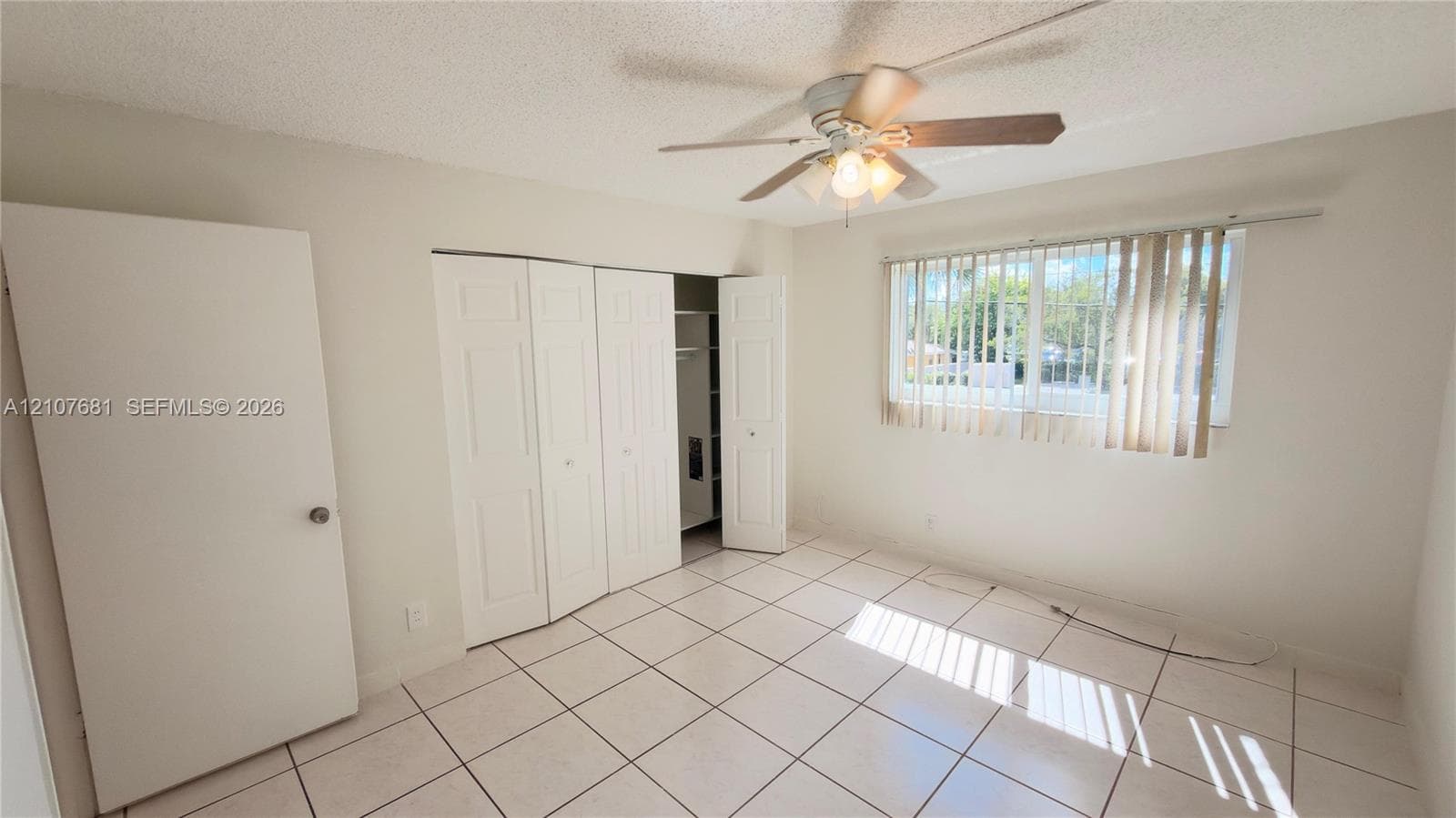 Room featuring white tile flooring, multi-panel bi-fold closet doors, ceiling fan with integrated lighting, and vertical window blinds