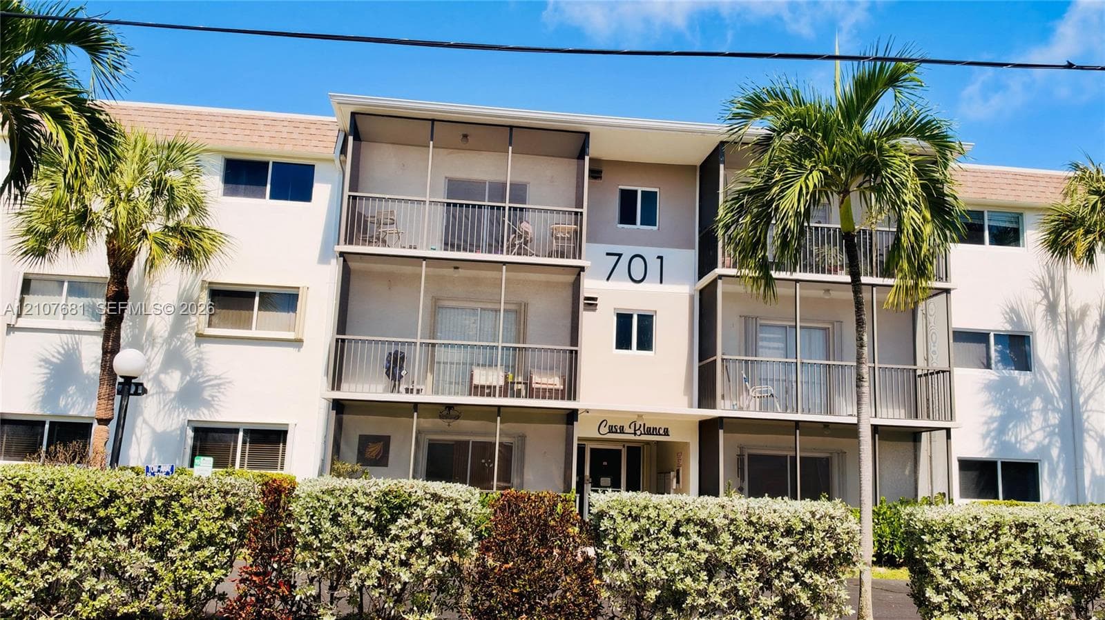 Multi-story stucco exterior featuring screened balconies, covered ground-level entry, and mature perimeter landscaping
