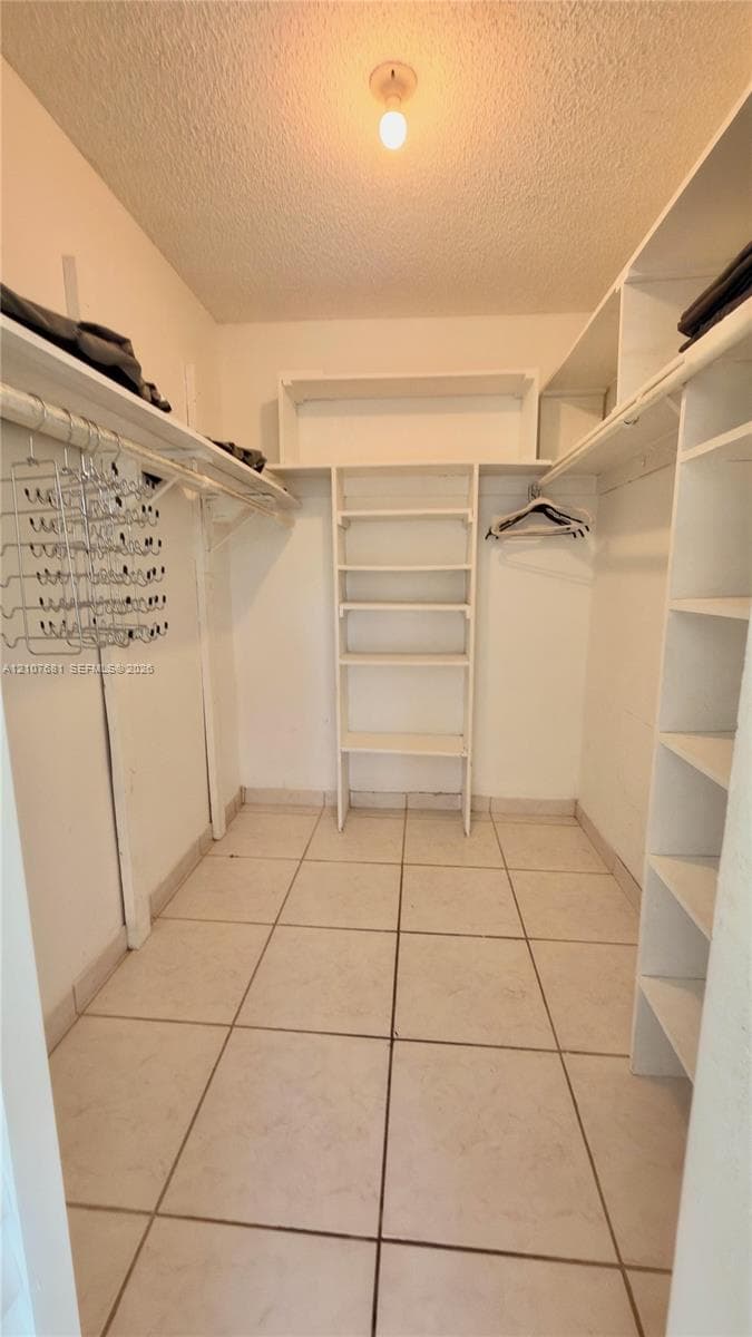 Walk-in closet featuring large-format tile flooring, built-in white shelving units, overhead hanging rods, and ceiling-mounted lighting