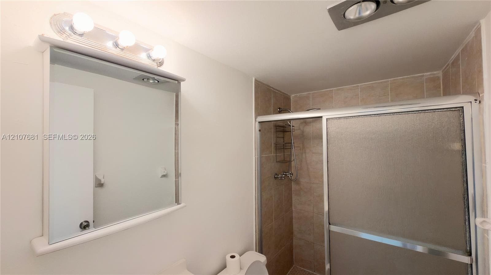 Bathroom featuring a shower stall with sliding glass doors, neutral-toned tiled walls, a wall-mounted vanity mirror with a three-bulb light fixture, and an integrated ceiling exhaust fan