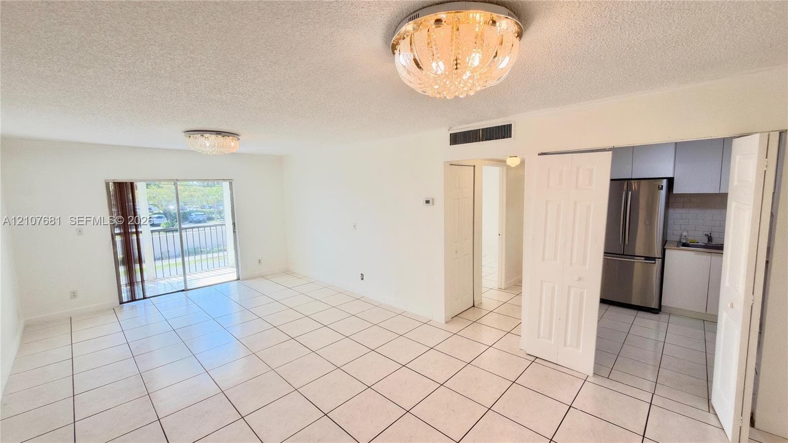 Open-concept area featuring white ceramic tile flooring, crystal-style flush-mount ceiling light fixtures, sliding glass doors with balcony access, and a kitchen equipped with stainless steel appliances