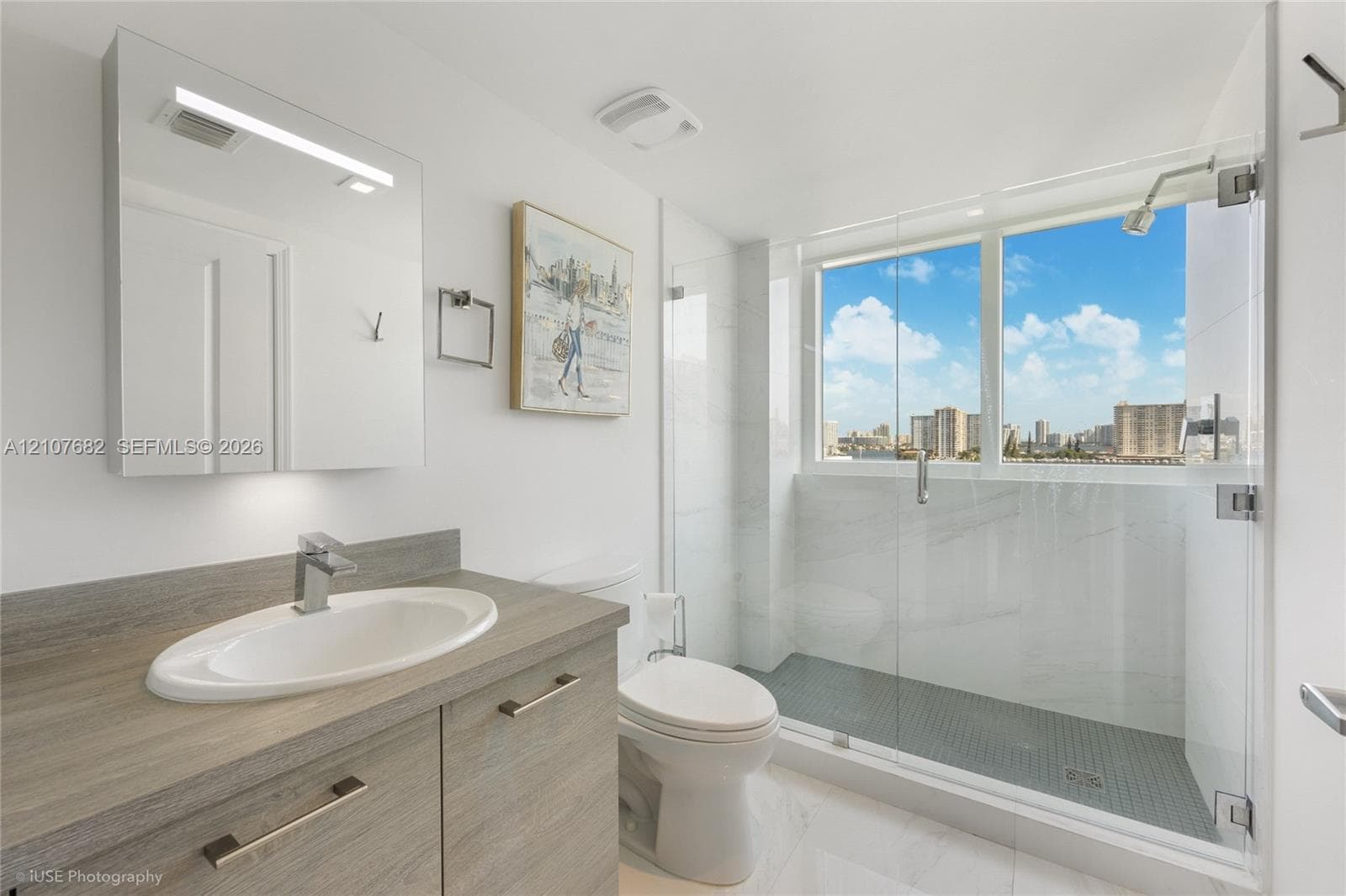 Bathroom featuring a wood-finish vanity with an integrated oval sink, glass-enclosed shower with tiled surround, and large-scale window