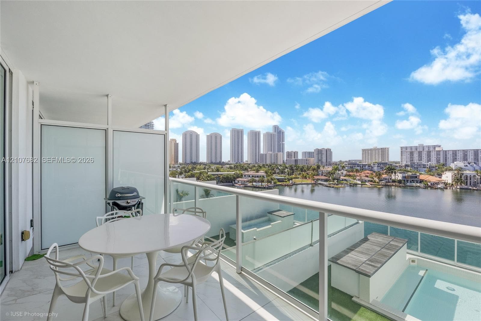 Covered balcony featuring stone-finish flooring, glass panel railing, and frosted privacy partitions, overlooking a waterfront skyline