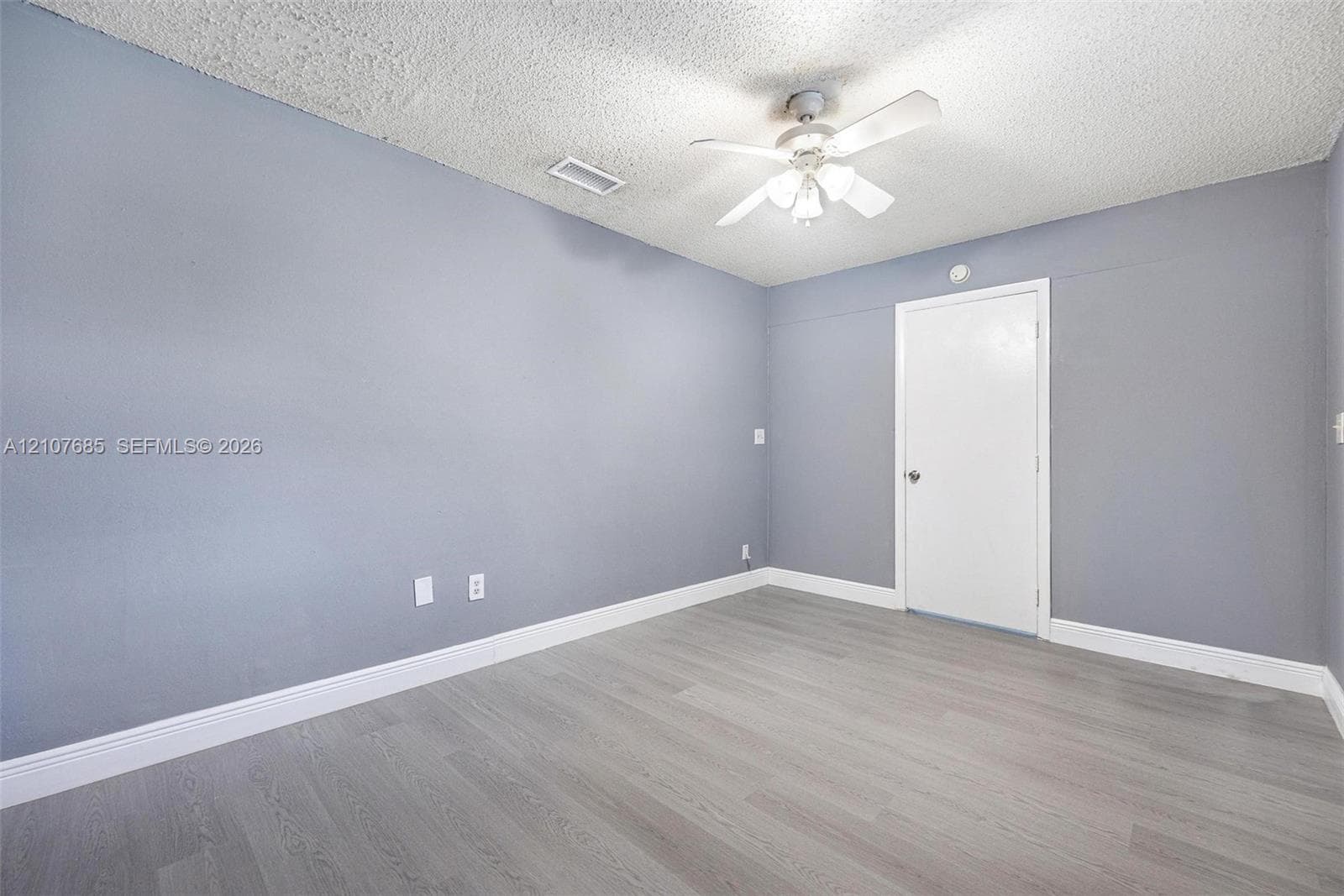 Neutral-toned interior room featuring wood-finish flooring, a ceiling-mounted light fixture with fan blades, and a solid-core interior door