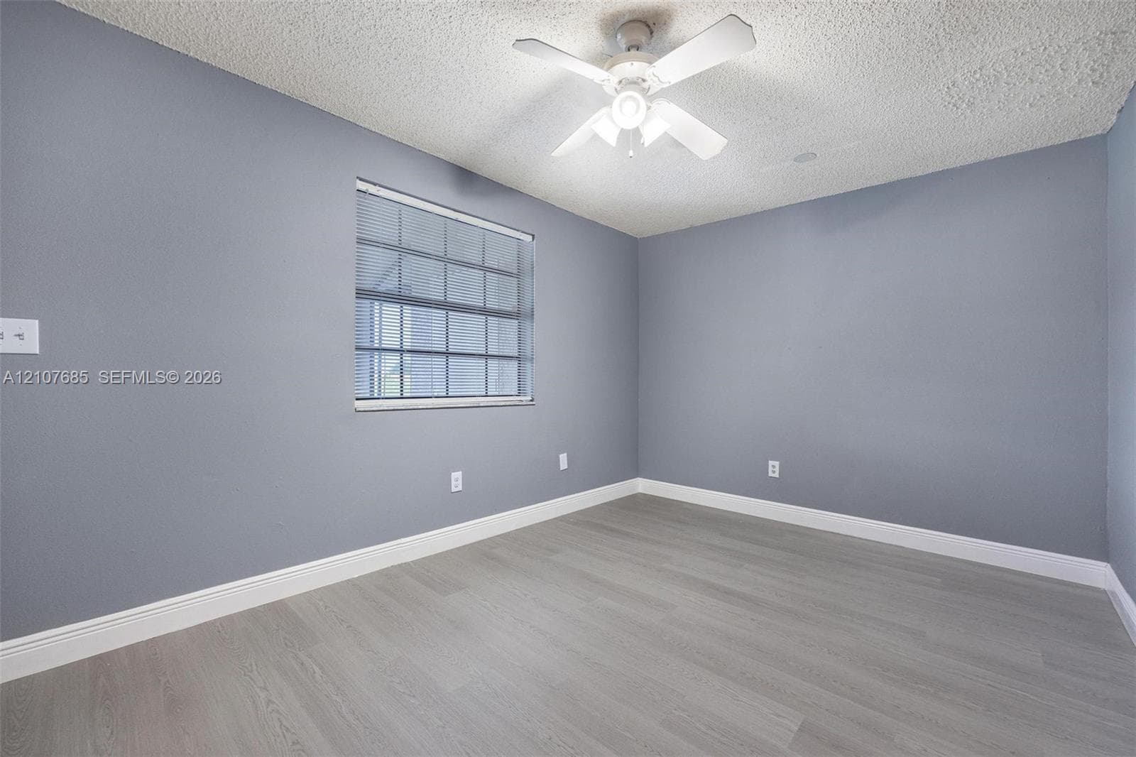 Room featuring wood-finish flooring, textured ceiling, light-toned wall paint, white-framed window with blinds, and a white ceiling fan with integrated light fixture