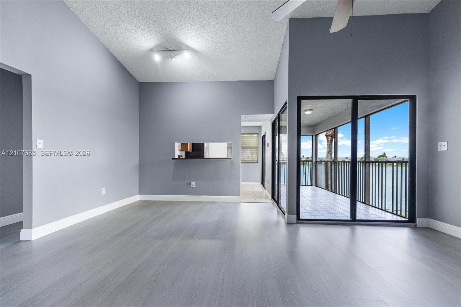Vaulted ceilings featuring a ceiling fan, neutral wall paint, wood-finish flooring, a kitchen pass-through window, and floor-to-ceiling glass sliding doors opening to a covered balcony