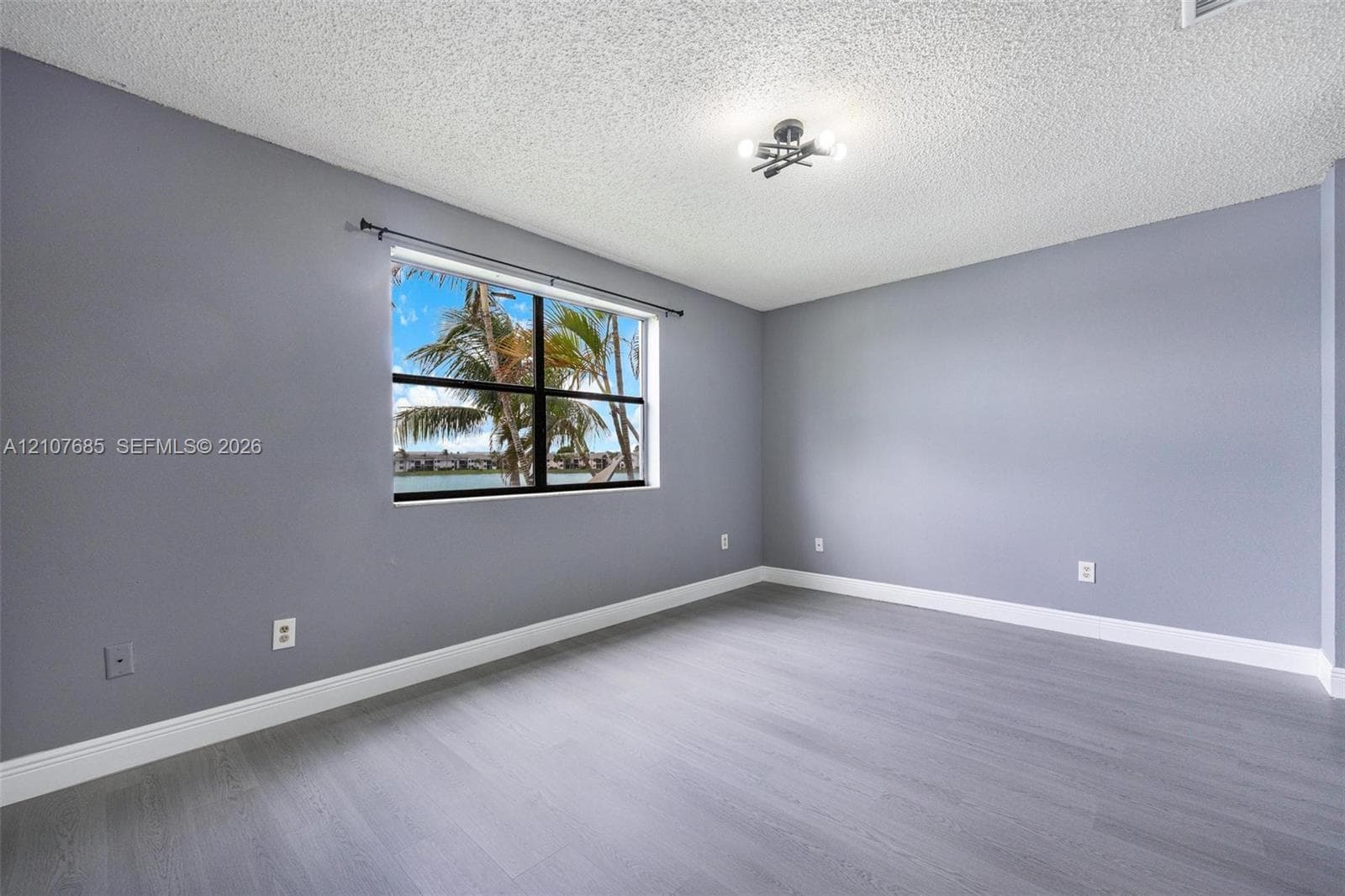 Room featuring gray-tone walls, wood-finish flooring, a multi-bulb flush-mount light fixture, and a single-pane window with exterior water and palm tree scenery