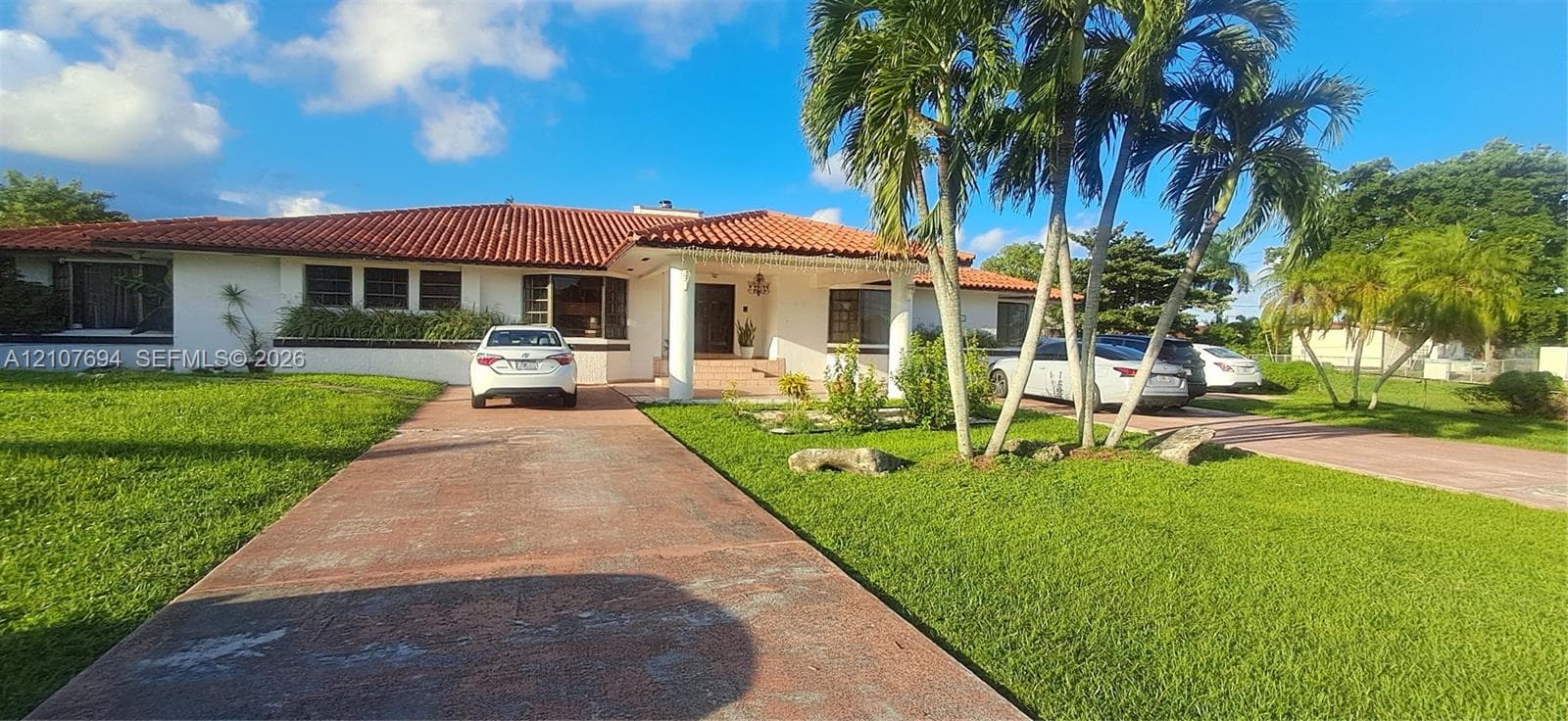 Single-story exterior featuring a terracotta tile roof, structural white columns, extensive paved driveway, and mature landscaping