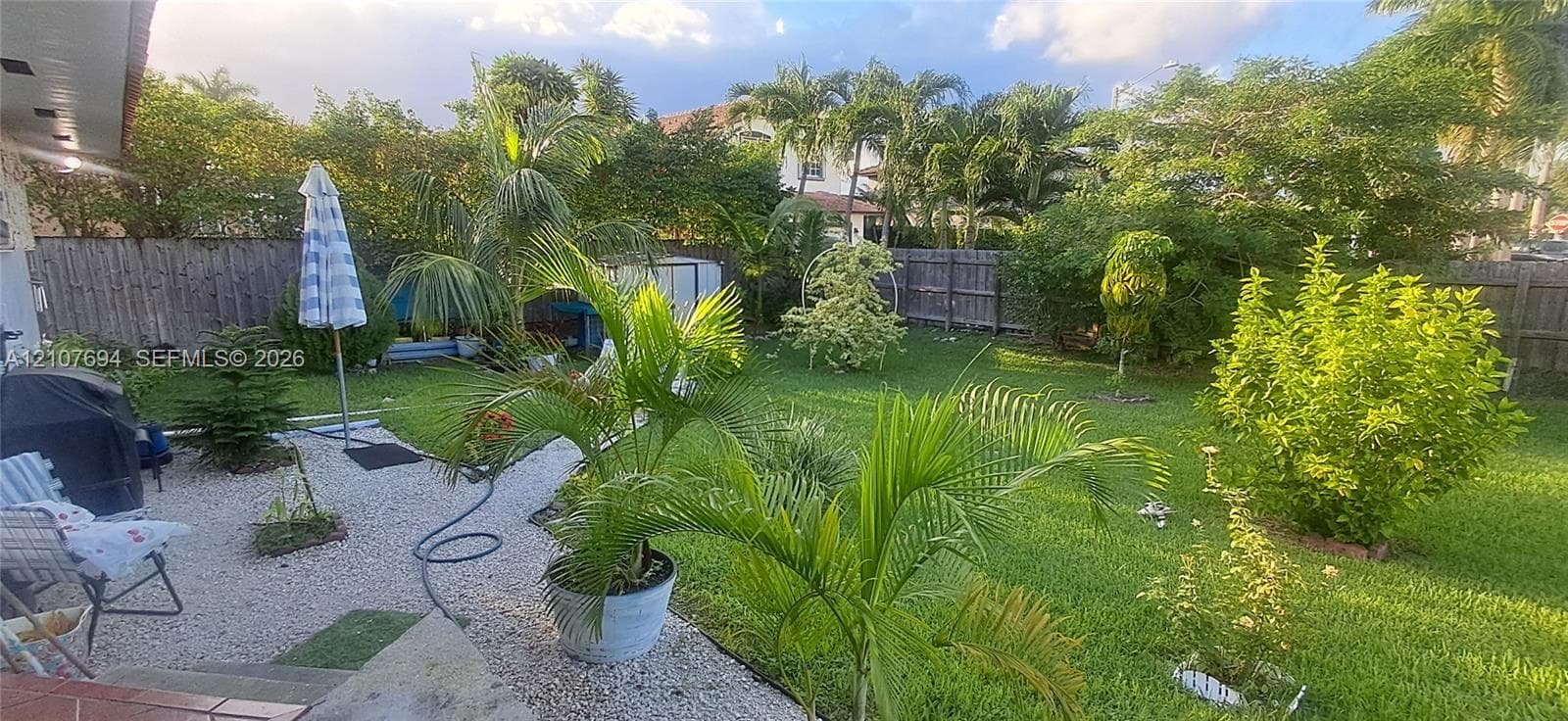 Backyard featuring a manicured lawn, stone-surfaced patio area, mature tropical landscaping, and perimeter wood fencing