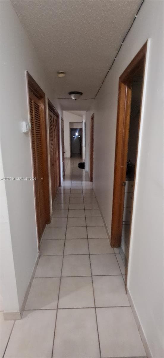 Hallway featuring neutral-toned tile flooring, louvered wood-finish closet doors, and flush-mount lighting fixtures
