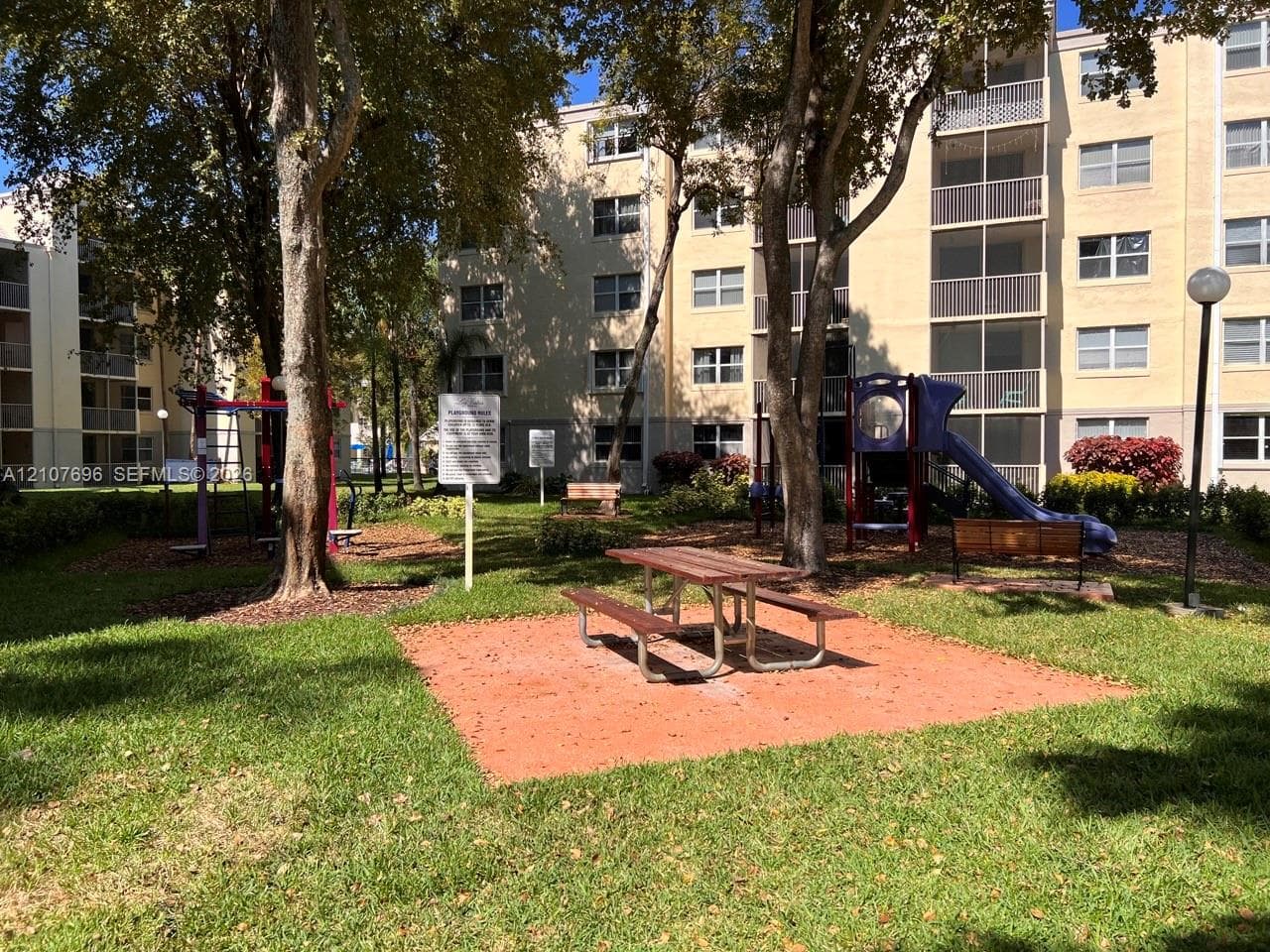 Common area featuring a recreational playground, manicured lawn, mature trees, and a fixed metal picnic table