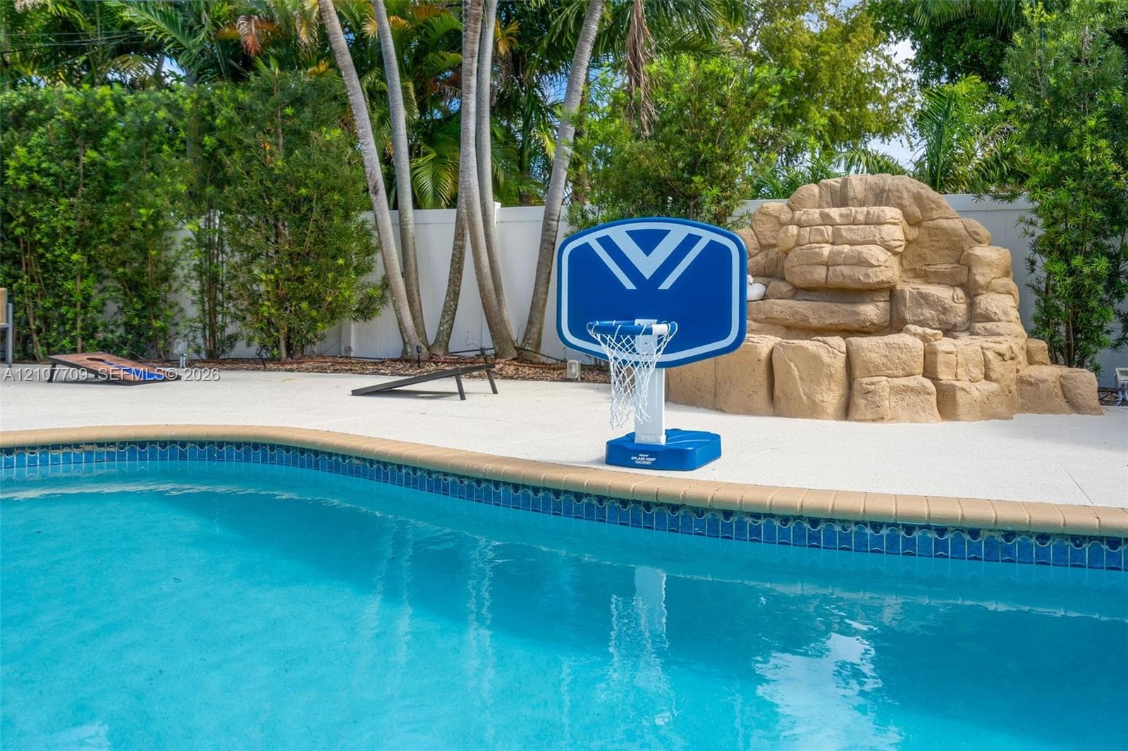 Swimming pool with tiled coping, decorative stone-finish waterfall feature, and concrete pool deck, bordered by mature palm trees and perimeter privacy fencing