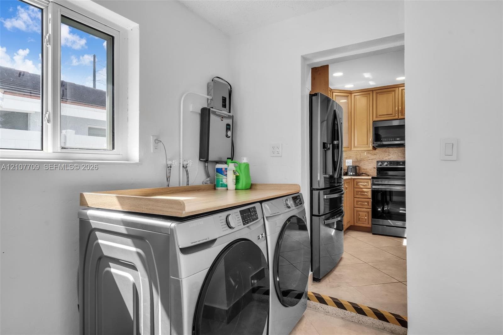 Laundry space featuring a wooden folding counter, vinyl-frame window, and tiled flooring extending into the kitchen area with wood-finish cabinetry and stainless steel appliances