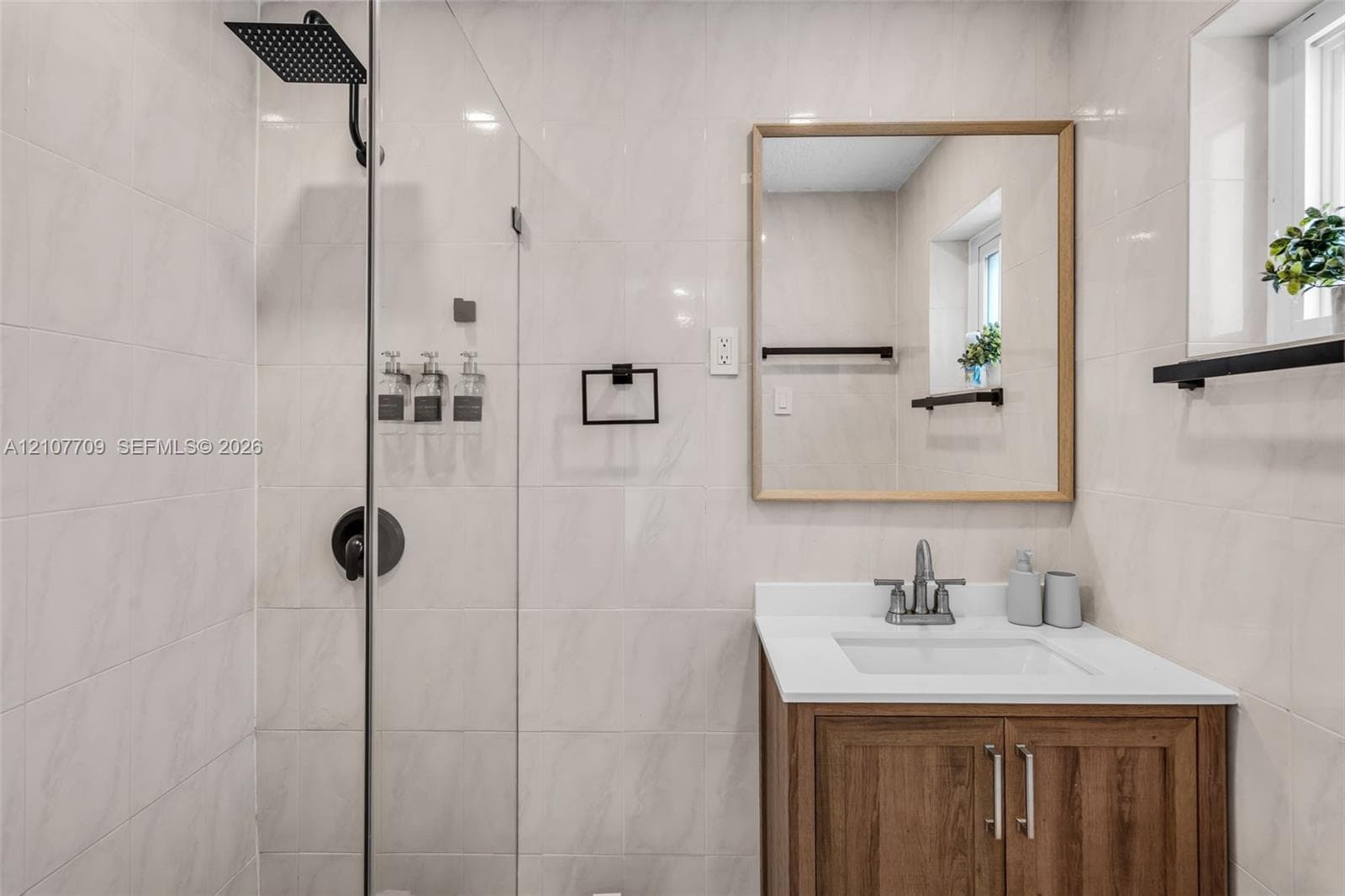 Bathroom featuring a glass-enclosed shower with matte black rainfall showerhead, wood-finish vanity with a white countertop, rectangular undermount sink, and wood-framed mirror