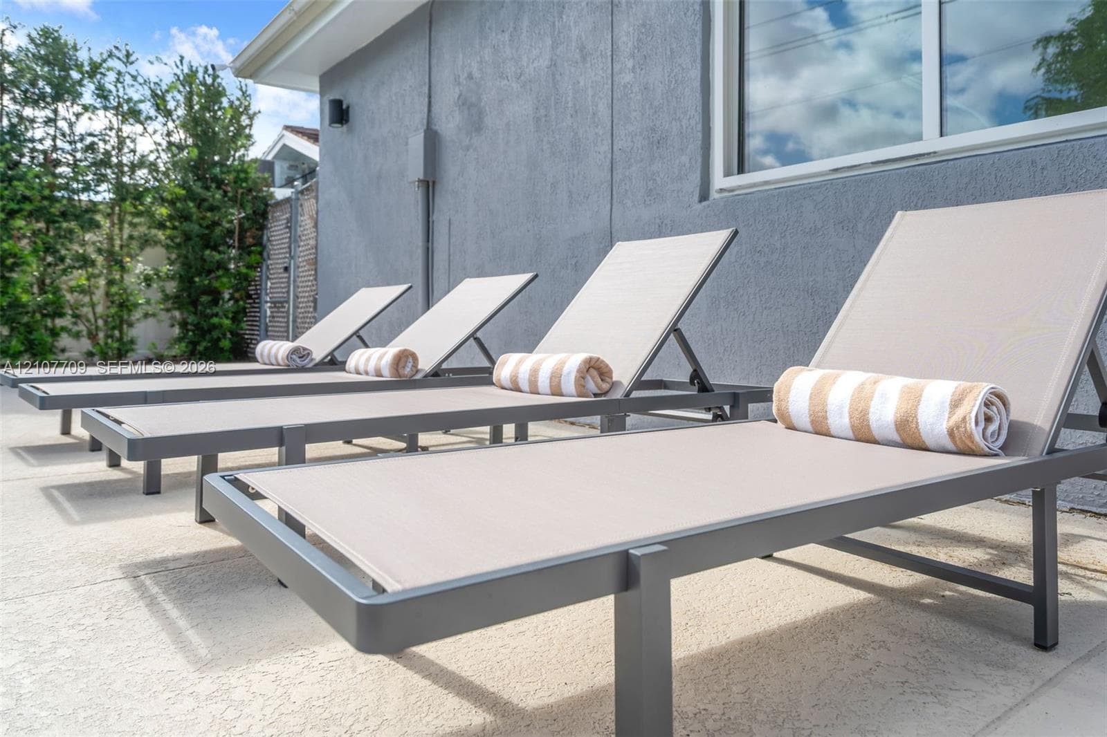 Textured gray stucco exterior featuring a concrete patio surface, white-trimmed window, and tall perimeter hedges