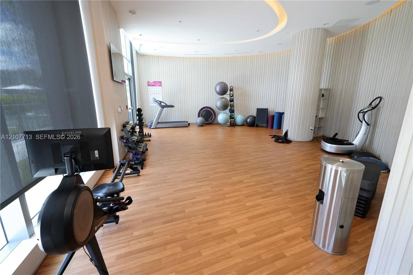 Fitness center featuring wood-finish flooring, curved walls with vertical paneling, recessed cove ceiling lighting, floor-to-ceiling windows, and a cylindrical support column