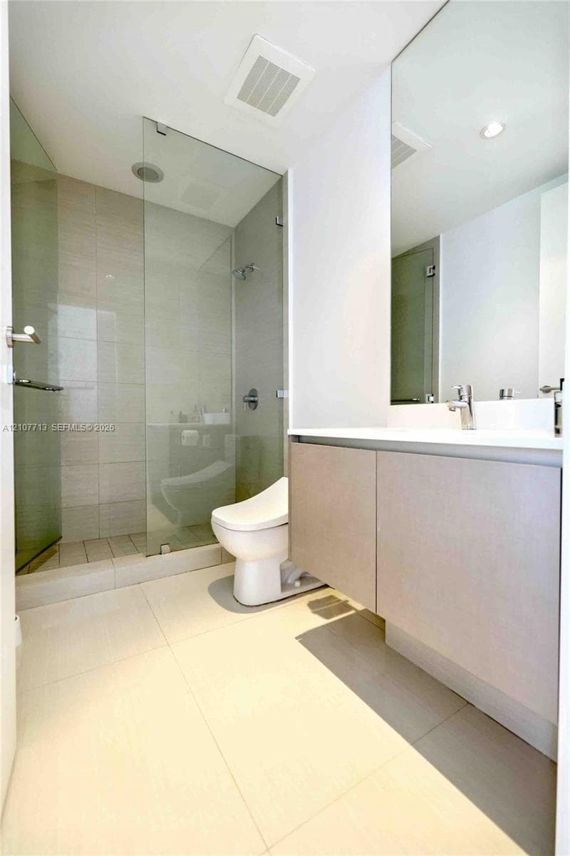 Floating vanity with solid surface countertop, frameless glass-enclosed shower featuring tile surrounds, wall-to-wall mirror, and large-format floor tiling