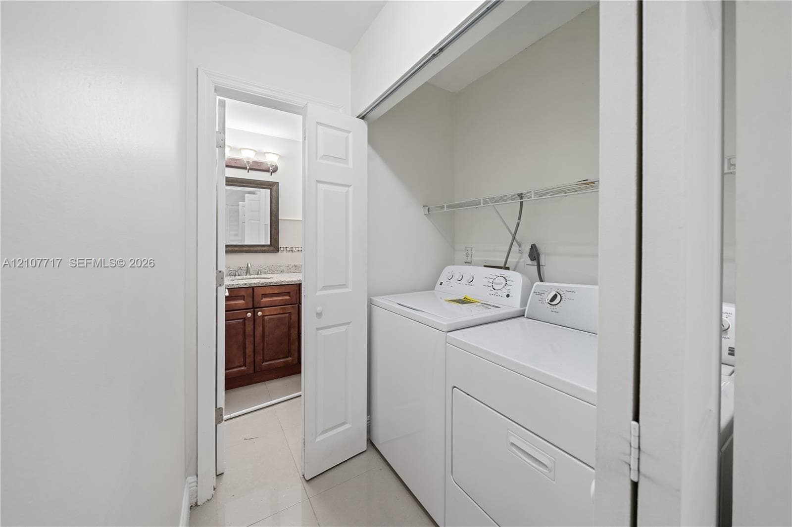 Laundry closet featuring white top-load appliances and a wire storage shelf, with an entry to a bathroom showcasing dark wood cabinetry, stone-finish countertops, and a wall-mounted light fixture