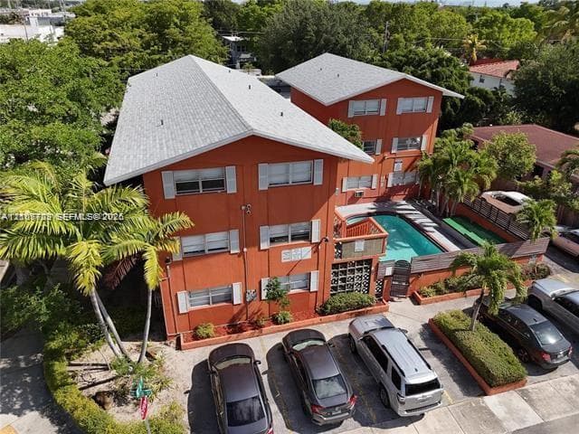Three-story multi-unit structure featuring a low-slope roof, exterior swimming pool, surface parking, and mature palm trees