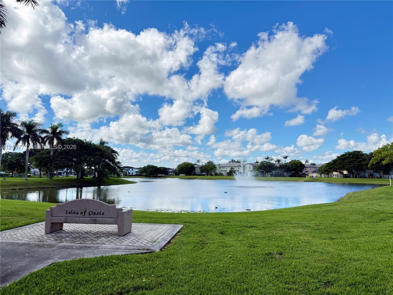 Community pond featuring a central fountain, manicured lawn, stone-finish bench, and patterned paver surround