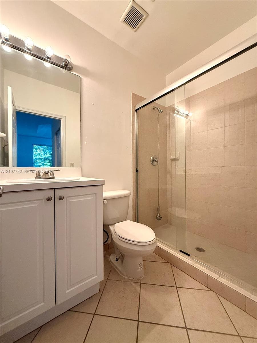 Bathroom featuring a white vanity cabinet, rectangular mirror with an attached four-bulb fixture, ceramic tile flooring, and a walk-in shower with a glass sliding door