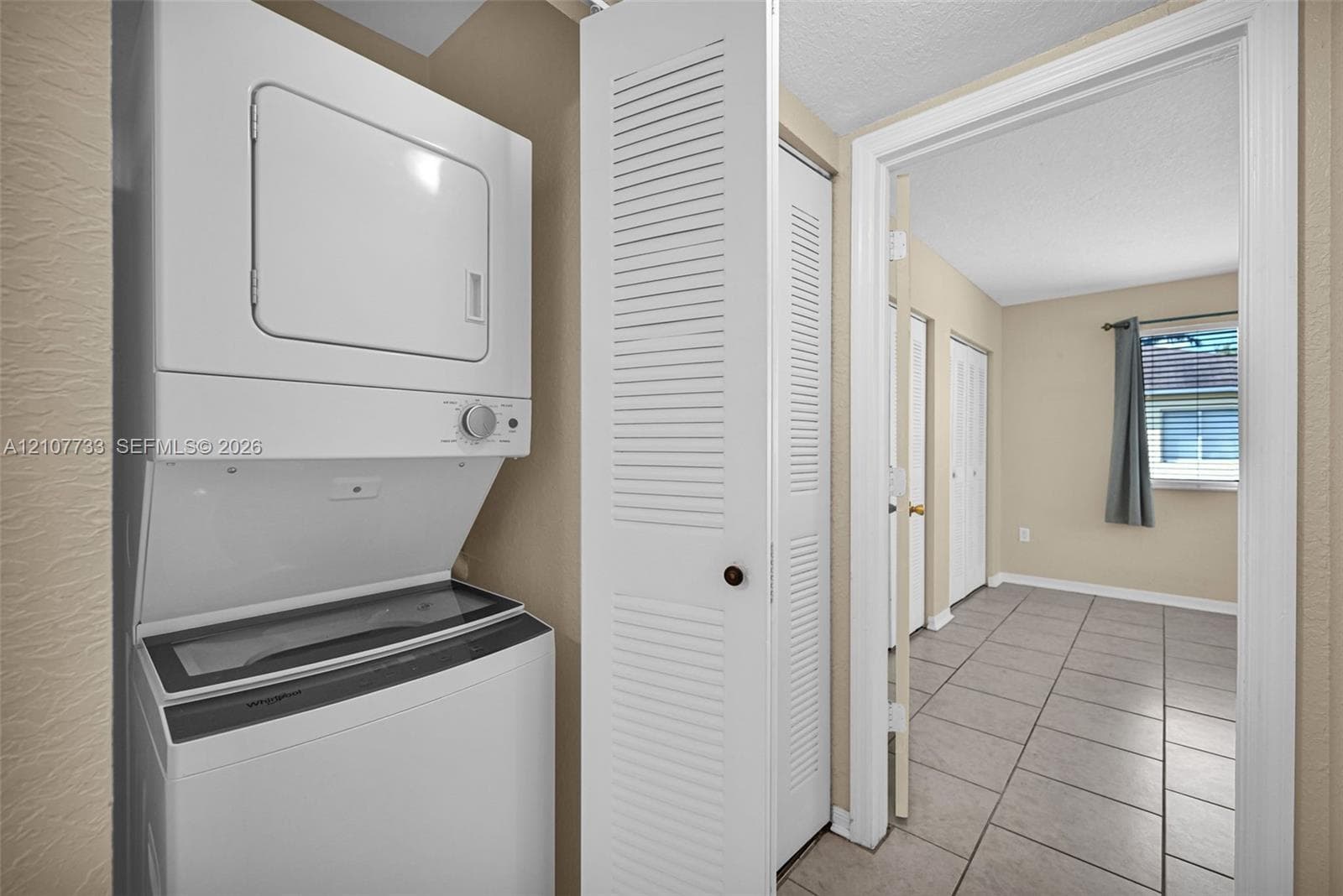 Laundry area featuring a stacked washer and dryer unit, white louvered bi-fold closet doors, and ceramic tile flooring