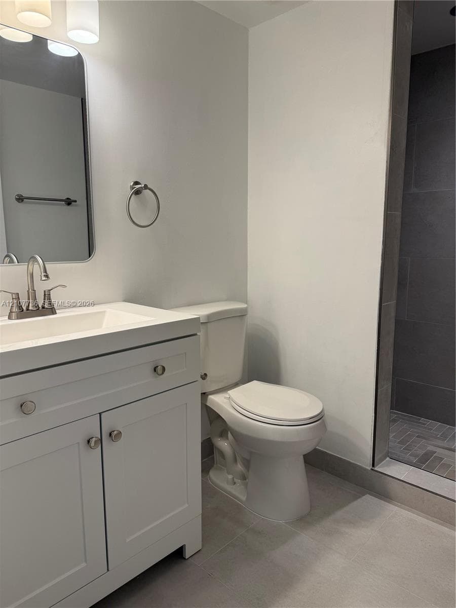 White vanity with integrated rectangular sink and metallic hardware, rounded rectangular mirror, three-light overhead fixture, and shower with dark tile surround and herringbone-patterned floor tile