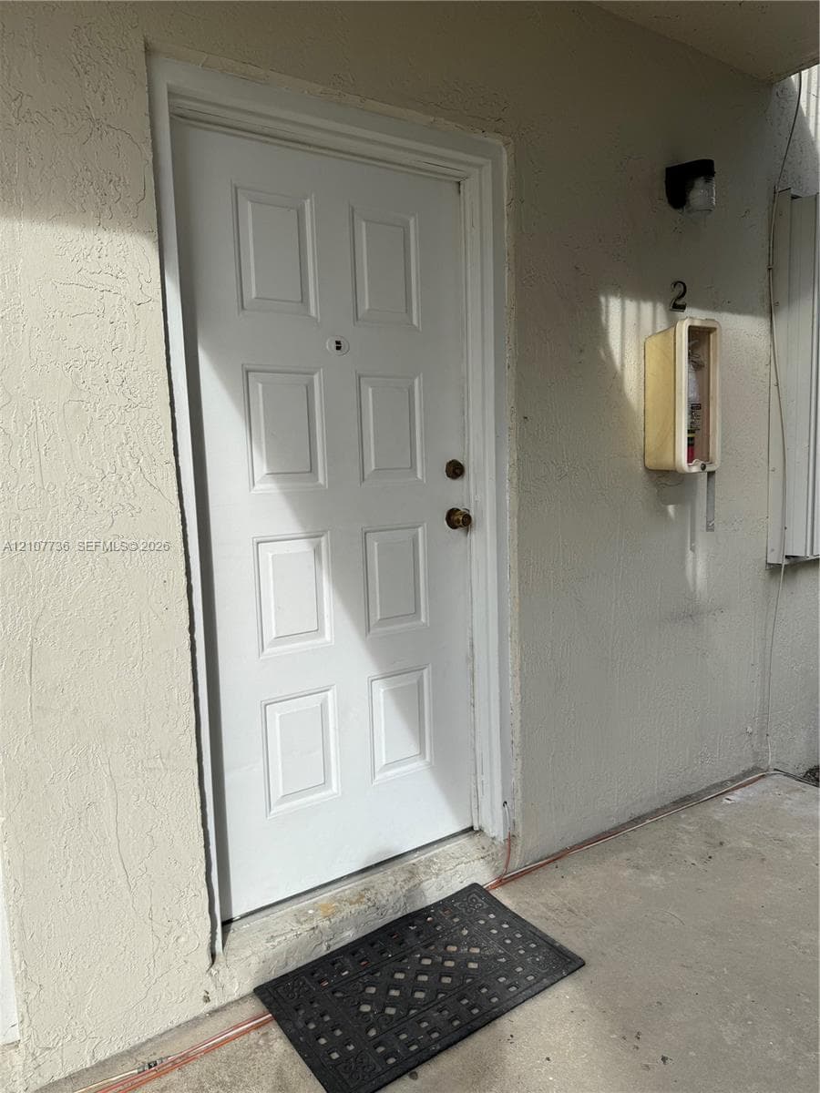 White six-panel entry door featuring a central peephole and brass-finish hardware, set within a stucco exterior wall