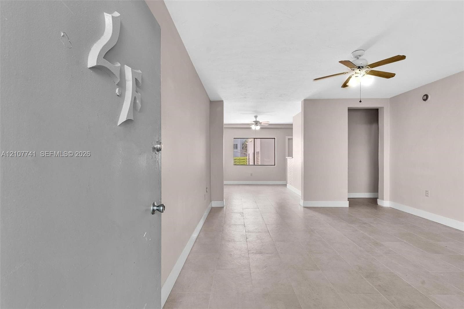 Interior space featuring large-format neutral tile flooring, multi-pane window with dark framing, wall-mounted air conditioning unit, and a ceiling-mounted fan with integrated lighting