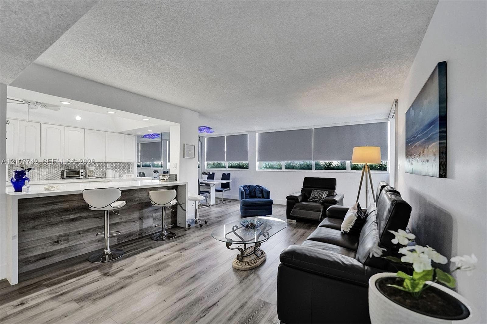 Open-concept interior featuring wood-finish flooring, a kitchen peninsula with bar seating, white cabinetry, a mosaic backsplash, and expansive windows