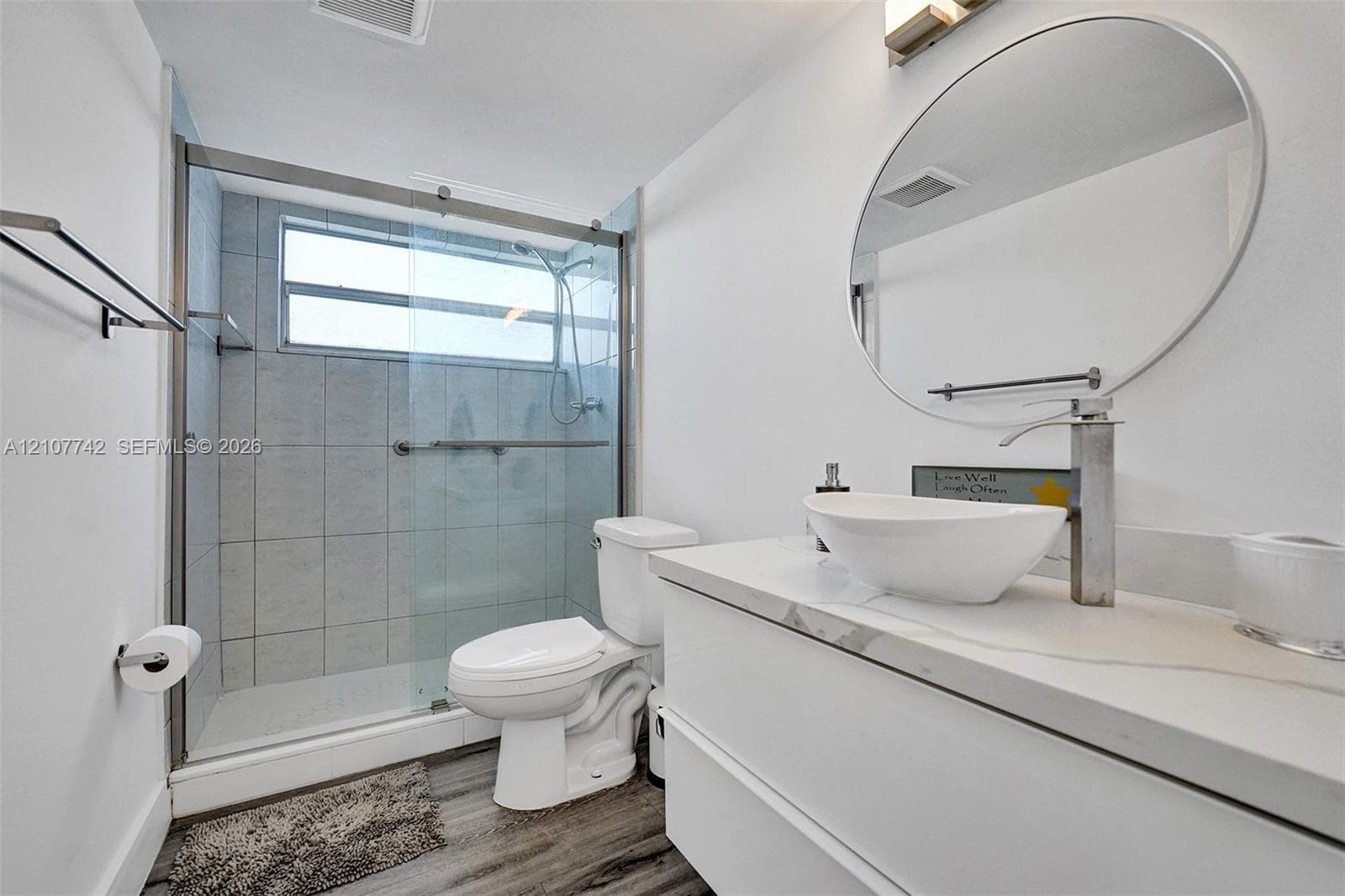 Modern bathroom featuring a floating vanity with a white countertop and vessel sink, circular mirror, and walk-in shower with a glass enclosure and tiled surround