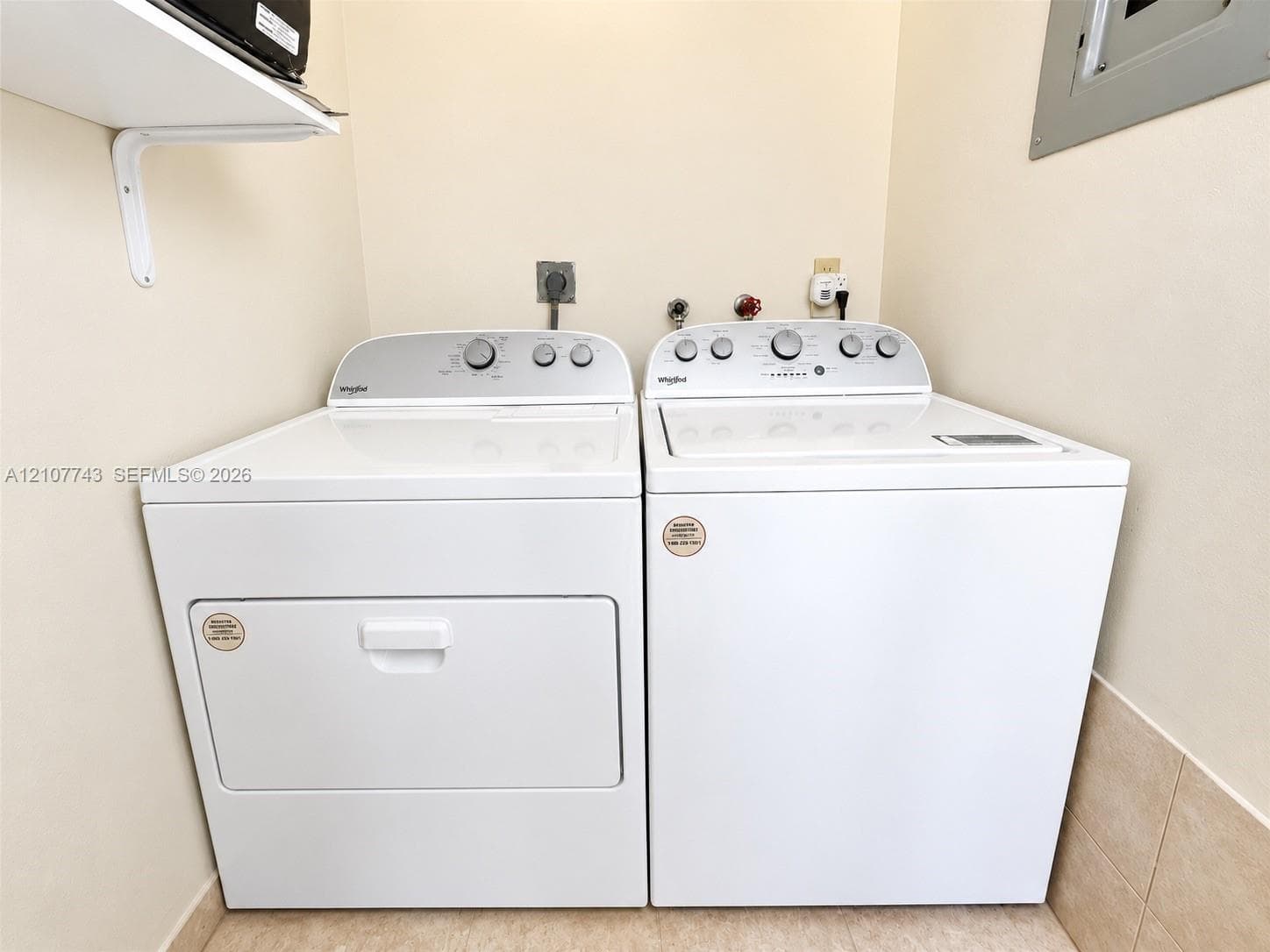 Laundry area featuring white top-load washer and dryer set, neutral tile flooring, and overhead wall-mounted shelving