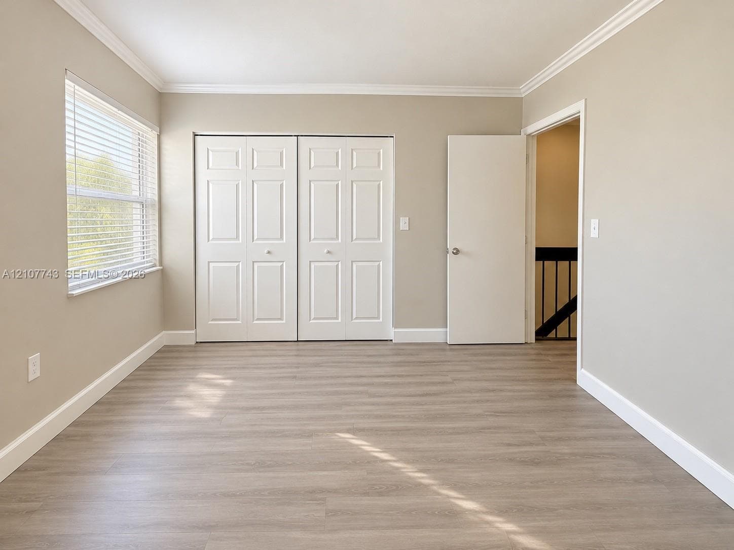 Neutral-toned interior featuring wood-finish flooring, crown molding, double-panel closet doors, and a window with white horizontal blinds