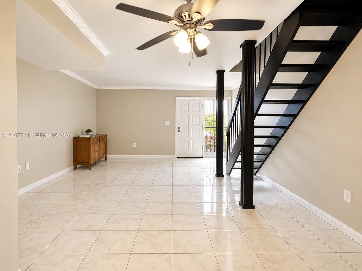 Neutral-toned ceramic tile flooring, open-tread staircase with dark-finished railings, fluted support column, ceiling-mounted fan with light fixture, and crown molding
