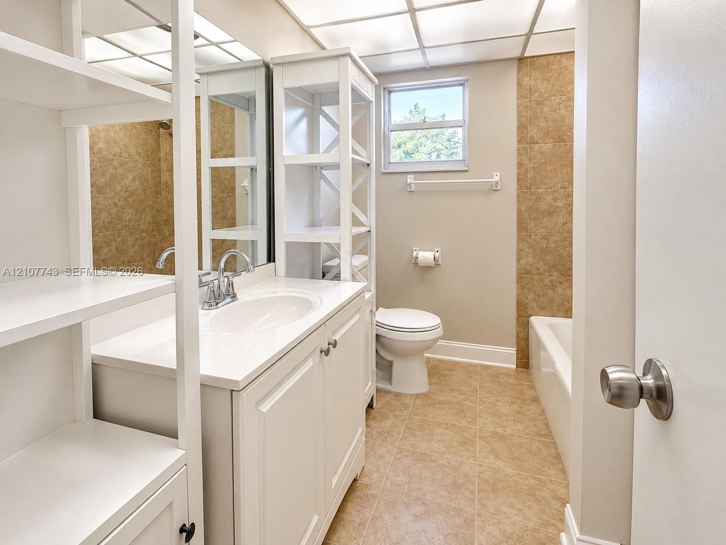 Bathroom featuring a white vanity with an integrated basin, built-in shelving unit, tile-surround bathtub, and neutral stone-finish flooring
