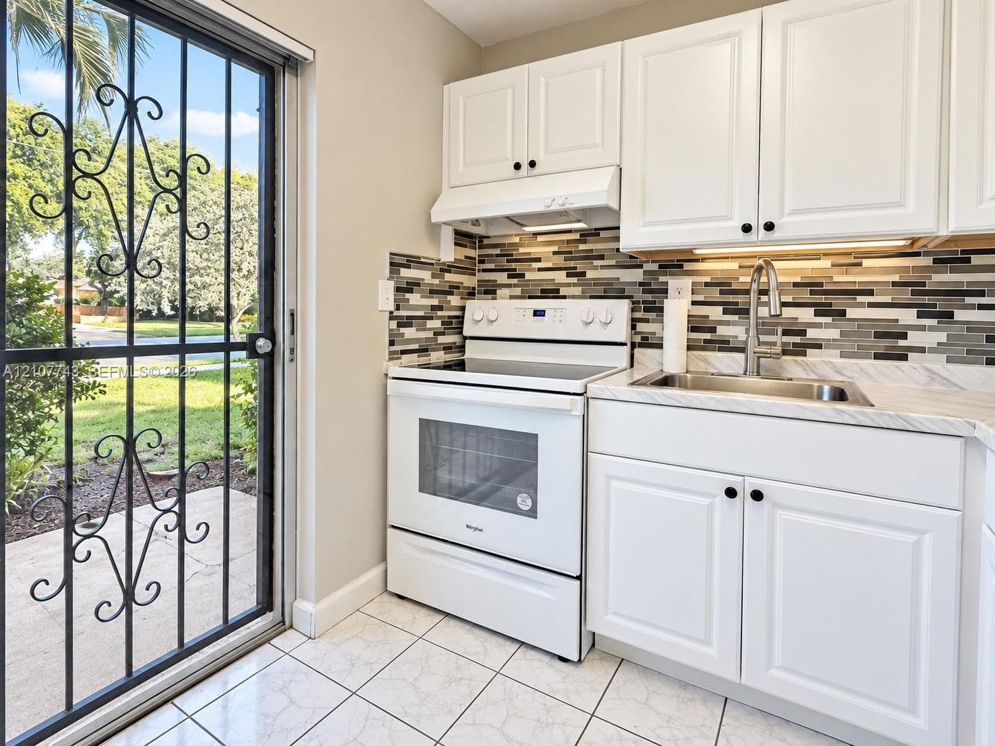 Kitchen featuring white cabinetry, a multi-tone tile backsplash, stone-finish countertops, stainless steel sink with a high-arch faucet, and white tile flooring