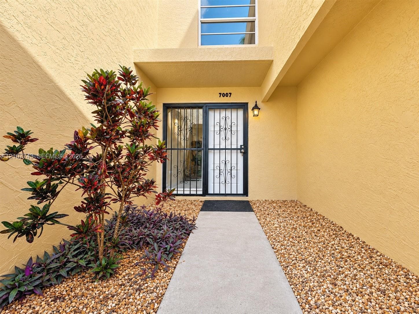 Entrance featuring a concrete walkway, decorative metal security door, stucco exterior, and rock-based landscaping with ornamental plantings