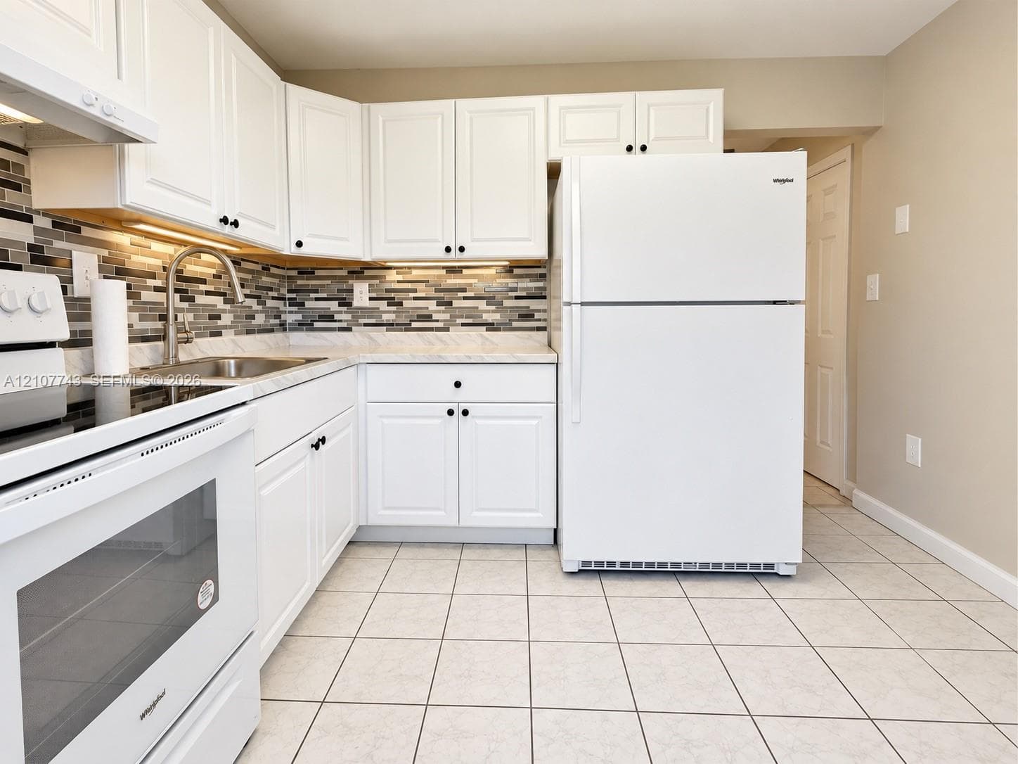 Kitchen featuring white shaker-style cabinetry, mosaic tile backsplash, stone-finish countertops, stainless steel undermount sink with arched faucet, and tiled flooring