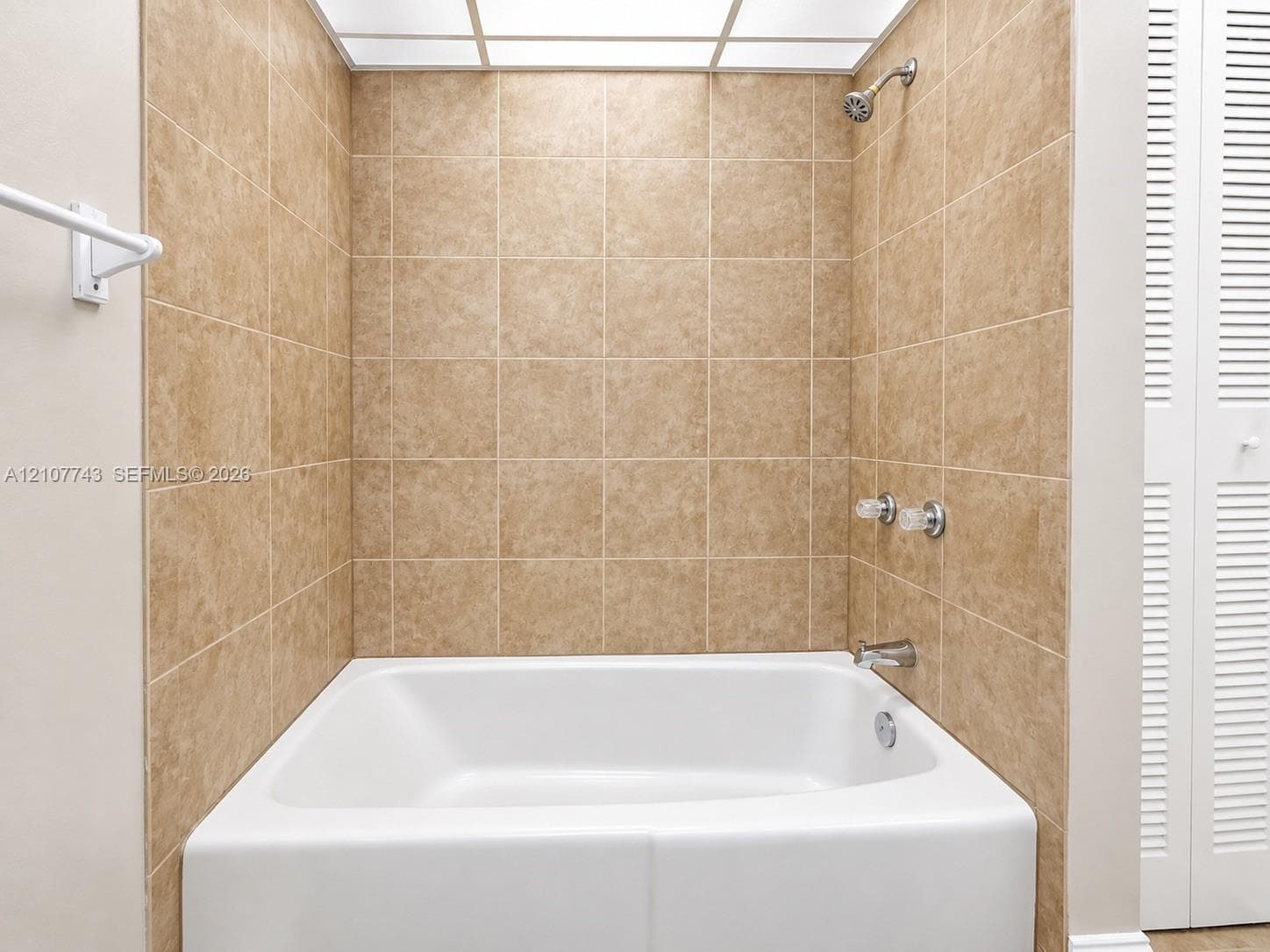 Bathtub alcove featuring beige tile surround, integrated shower head, chrome-finish faucet handles, and a grid-style ceiling light fixture