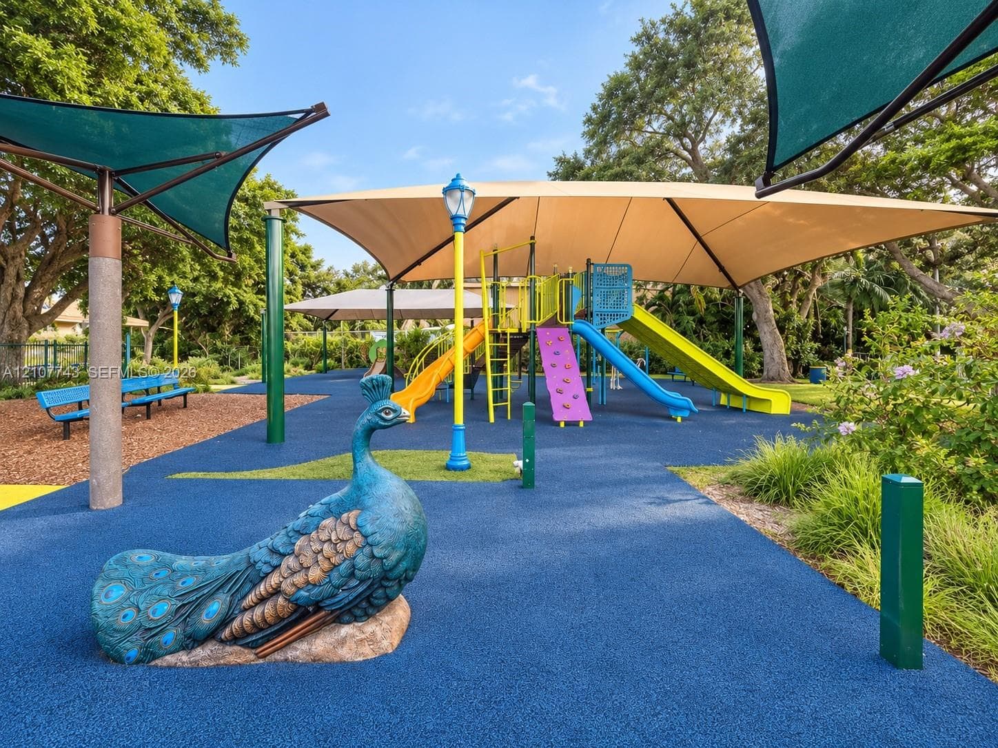 Recreational area featuring a playground structure with multiple slides and a climbing wall, sheltered by overhead fabric shade canopies, finished with blue poured-rubber surfacing, and accented by a decorative peacock statue
