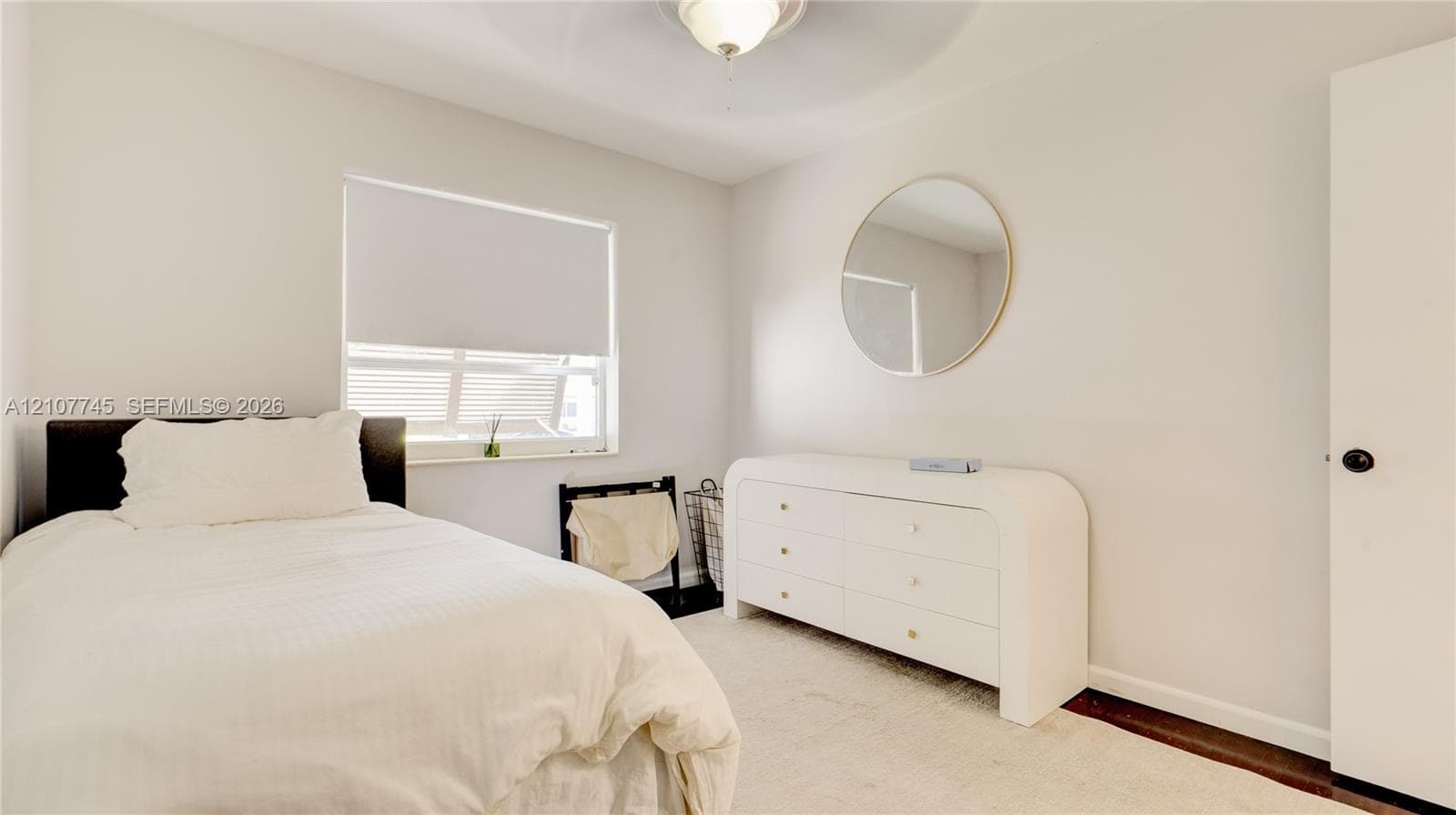 Neutral wall palette, carpeted flooring, circular wall-mounted mirror, white window roller shade, and ceiling-mounted light fixture