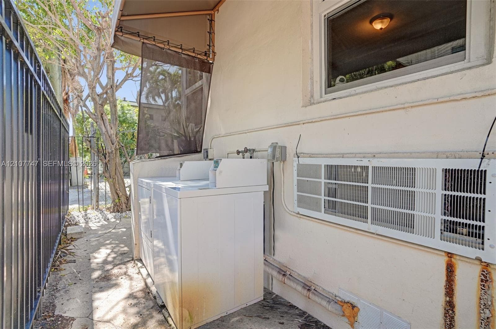 Exterior utility area featuring a wall-mounted ventilation unit, a metal fence enclosure, and a built-in awning