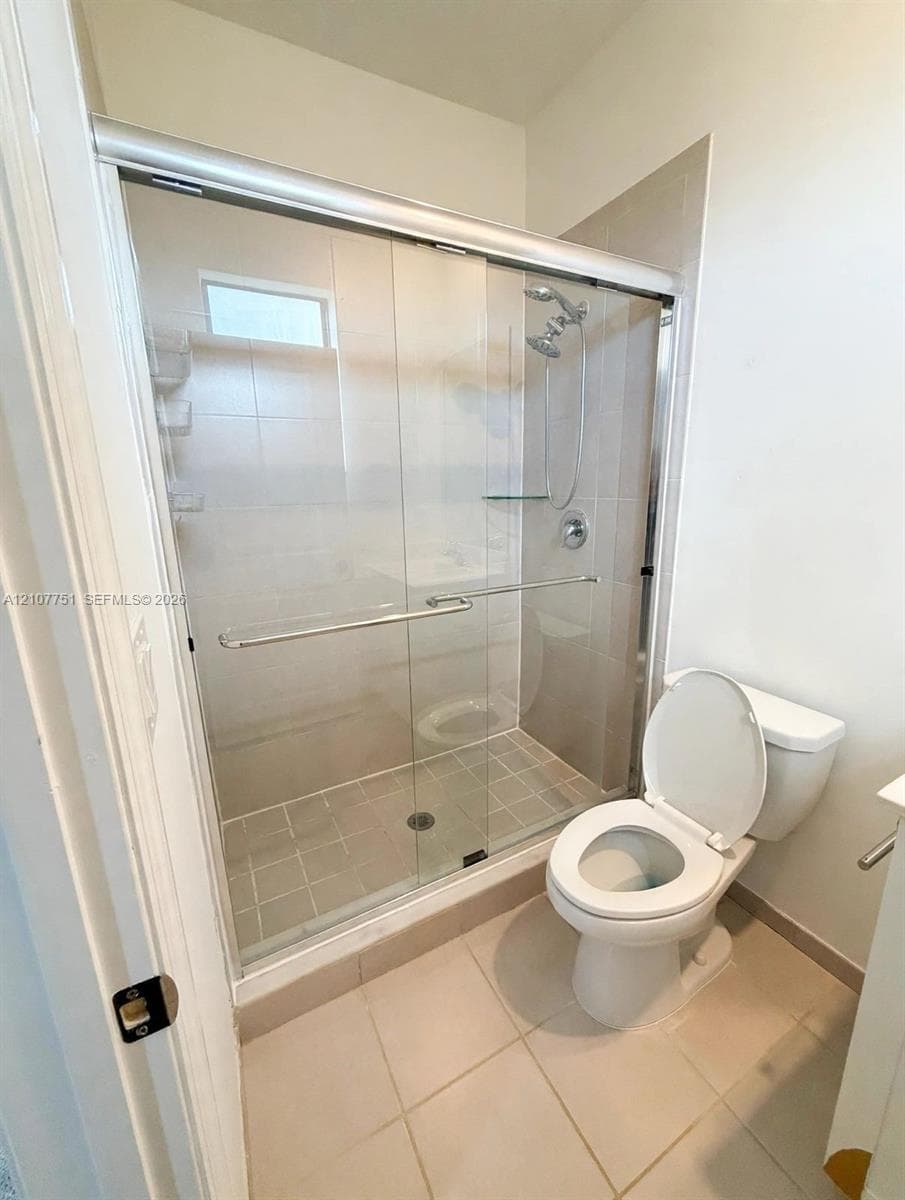 Walk-in shower featuring a sliding glass enclosure, light-toned wall tile, and an adjustable handheld showerhead, accompanied by a ceramic toilet and large-format floor tile