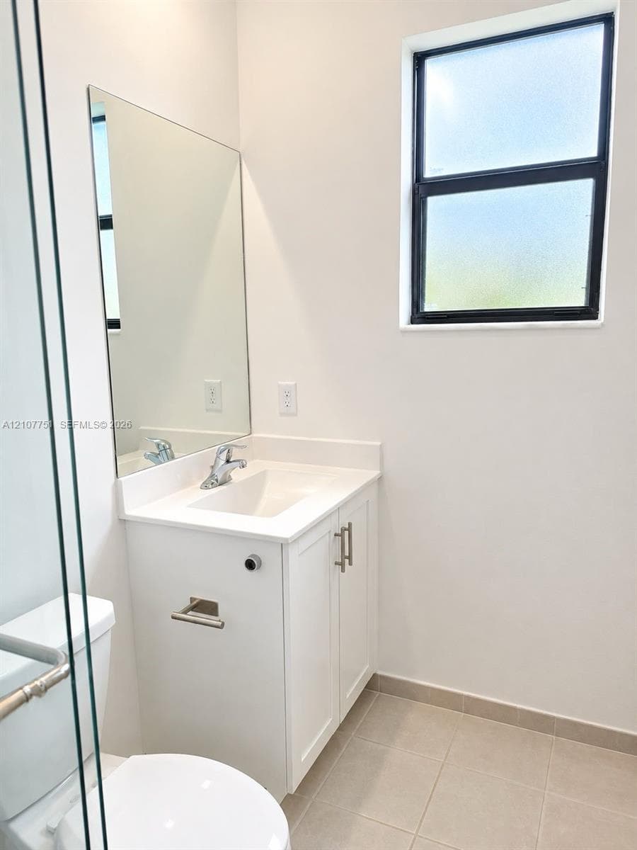 Bathroom featuring a white vanity cabinet with an integrated rectangular sink, solid-surface countertop, frameless wall-mounted mirror, and a window with a black frame