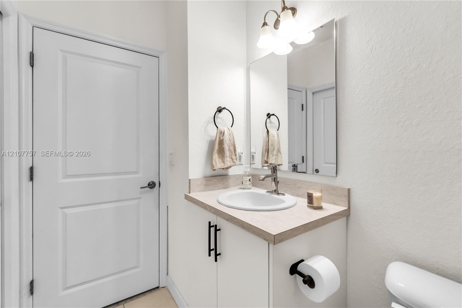 Vanity featuring a light-toned countertop, undermount oval sink, matte black hardware, and an attached three-light fixture above a rectangular mirror